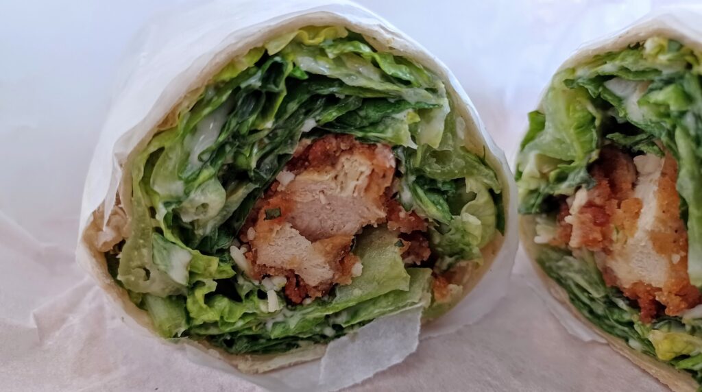 Ultra close-up macro cross-section of a sliced crispy chicken Caesar wrap revealing golden crunchy chicken tenders, crisp romaine lettuce, shaved parmesan, and creamy garlic dressing